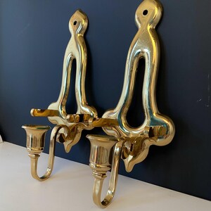 May include: Two ornate, gold-colored wall sconces. Each sconce features a tall, curved backplate with a decorative top and a candle holder. The sconces are made of a shiny, reflective metal and are mounted on a dark blue wall.