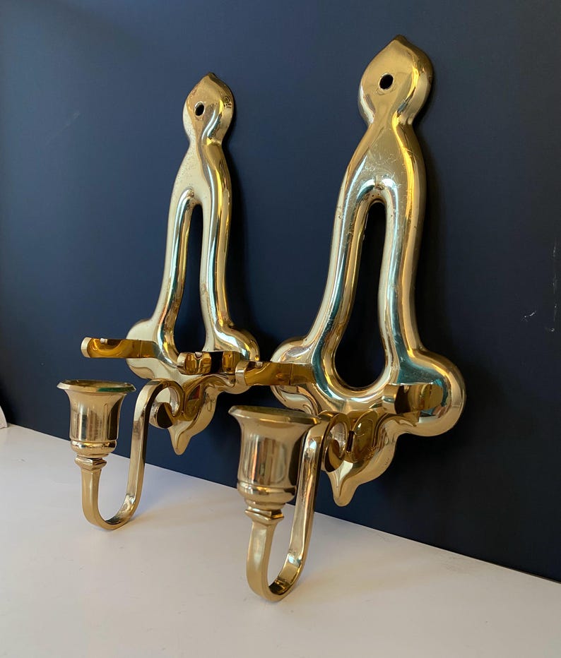May include: Two ornate, gold-colored wall sconces. Each sconce features a tall, curved backplate with a decorative top and a candle holder. The sconces are made of a shiny, reflective metal and are mounted on a dark blue wall.