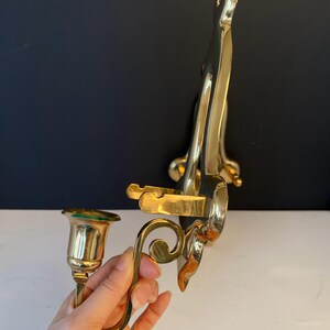 May include: A pair of brass wall sconces with a polished finish. One sconce holds a candle cup, and the other has a decorative, curved design. The sconces are designed to be mounted on a wall and add a touch of elegance.