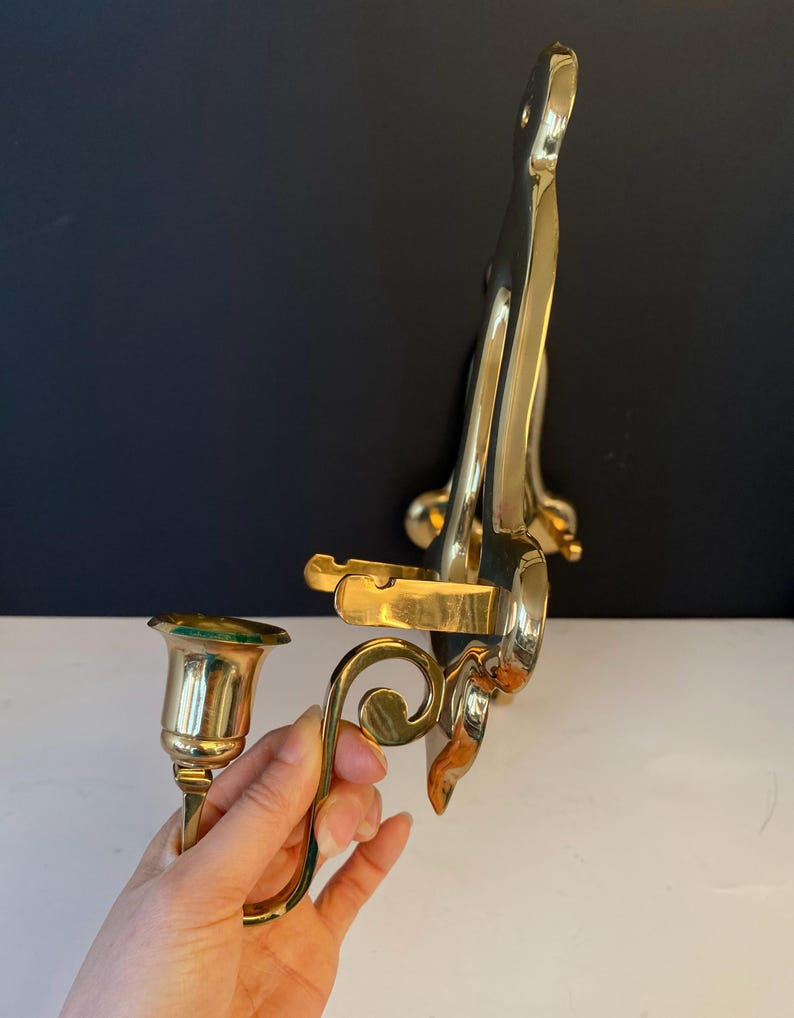 May include: A pair of brass wall sconces with a polished finish. One sconce holds a candle cup, and the other has a decorative, curved design. The sconces are designed to be mounted on a wall and add a touch of elegance.