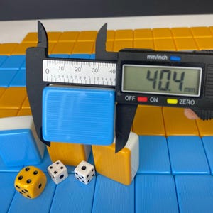 Puede incluir: Una ficha azul de un juego de Mahjong se mide con un calibrador digital, mostrando un ancho de 40,4 mm. La imagen tambi&eacute;n muestra otras fichas en azul, blanco y amarillo, junto con tres dados. Las fichas est&aacute;n dispuestas sobre una superficie azul y amarilla.