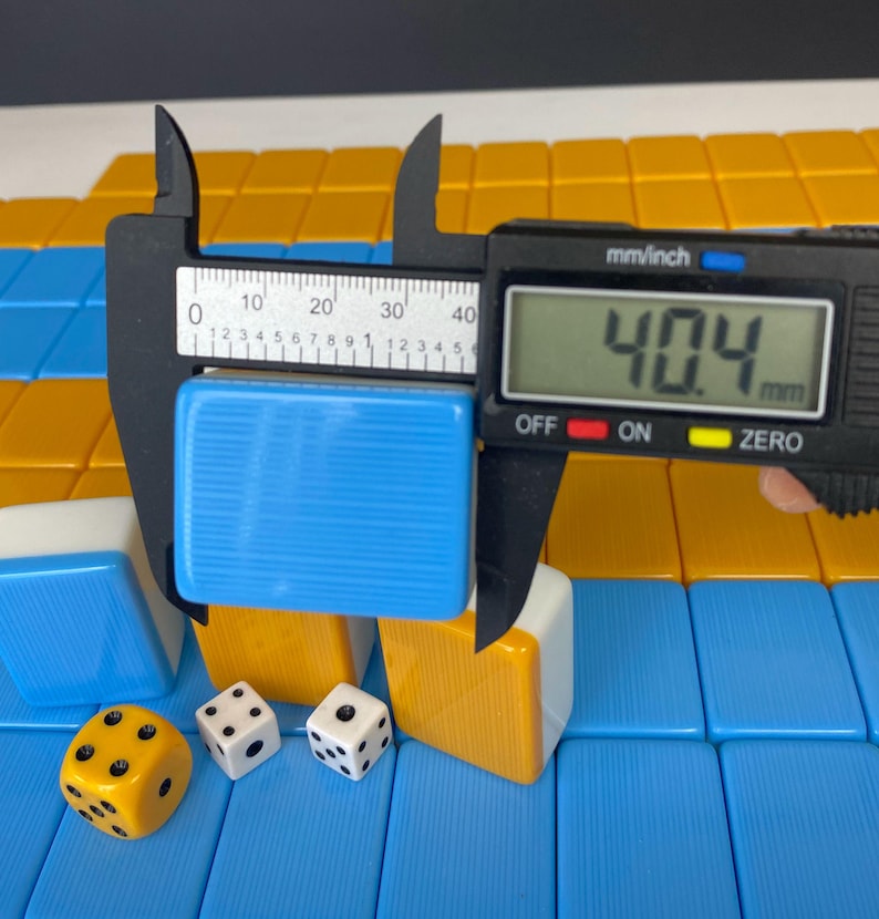Puede incluir: Una ficha azul de un juego de Mahjong se mide con un calibrador digital, mostrando un ancho de 40,4 mm. La imagen tambi&eacute;n muestra otras fichas en azul, blanco y amarillo, junto con tres dados. Las fichas est&aacute;n dispuestas sobre una superficie azul y amarilla.
