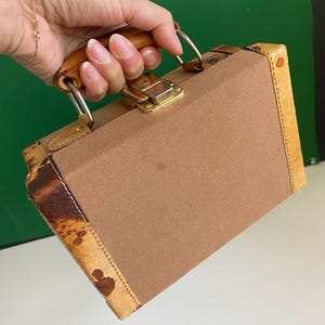 May include: A vintage brown suitcase with a tan canvas body, leather trim, and a wooden handle. The suitcase features a gold-colored clasp and metal hardware. The corners have a unique patterned design.