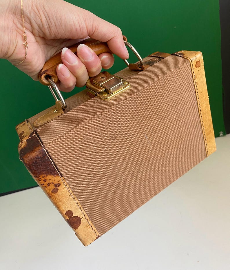 May include: A vintage brown suitcase with a tan canvas body, leather trim, and a wooden handle. The suitcase features a gold-colored clasp and metal hardware. The corners have a unique patterned design.