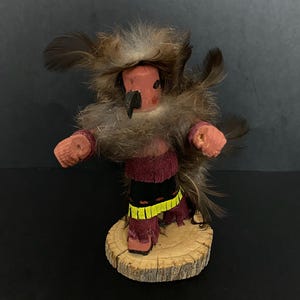 Può includere: Una bambola kachina nativa americana fatta a mano con un copricapo di piume, un viso dipinto di rosso e nero e una gonna viola e gialla. La bambola è in piedi su una base di legno.