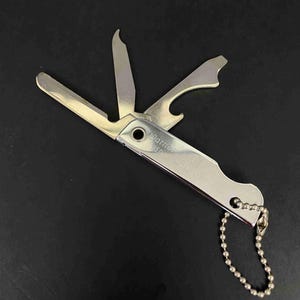 Peut inclure: Un outil multifonction argenté avec trois lames métalliques déployées, incluant un tournevis plat et un décapsuleur. Le manche porte l'inscription gravée Warner et RED, et une petite chaînette à billes en métal est fixée à l'extrémité.