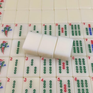 May include: A close-up of a Mahjong set with ivory-coloured tiles. Three tiles are in focus, with the numbers 3, 4, and 5 visible. The other tiles have green, red, and blue symbols and numbers.