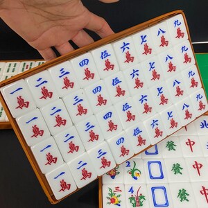 May include: A set of white Mahjong tiles with red and blue symbols and characters, arranged in a wooden box. The tiles are in a rectangular shape, with various numbers and suits. The game set is displayed on a dark surface.
