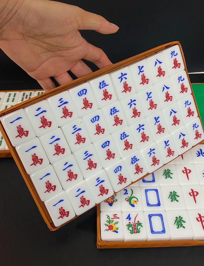 May include: A set of white Mahjong tiles with red and blue symbols and characters, arranged in a wooden box. The tiles are in a rectangular shape, with various numbers and suits. The game set is displayed on a dark surface.