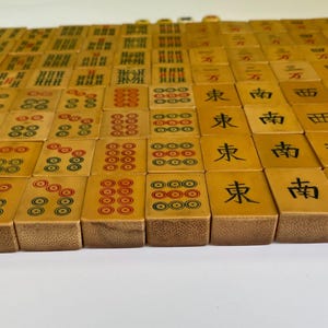 May include: A collection of vintage Mahjong tiles, each with unique markings and symbols in red, green, and black. The tiles are arranged in rows, showcasing the variety of designs and characters.