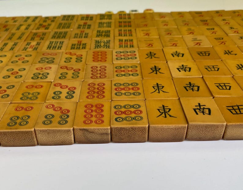 May include: A collection of vintage Mahjong tiles, each with unique markings and symbols in red, green, and black. The tiles are arranged in rows, showcasing the variety of designs and characters.