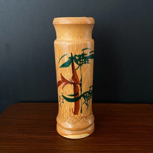 Puede incluir: Un jarrón de bambú marrón con un diseño pintado de tallos de bambú verdes y pinceladas rojas y negras. El jarrón tiene el texto "1974" y "Japón" escrito en él.