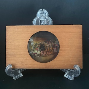 May include: A wooden frame with a circular historical image. The image shows a scene with several figures in period clothing. The frame is supported by clear, decorative stands, and the background is black.