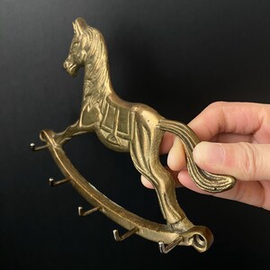 Könnte beinhalten: Ein Schlüsselhalter aus Messing in Form eines Schaukelpferdes mit fünf Haken. Das Pferd ist in Galopphaltung mit Sattel und Zaumzeug.