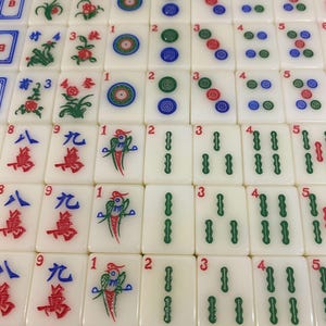 May include: A close-up of a set of ivory-coloured Mahjong tiles. The tiles feature various symbols, numbers, and colourful designs, including flowers, circles, and Chinese characters. The tiles are arranged in a grid pattern.