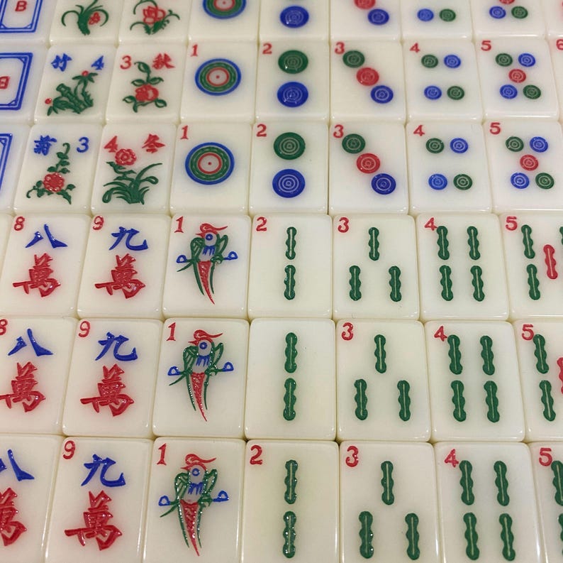 May include: A close-up of a set of ivory-coloured Mahjong tiles. The tiles feature various symbols, numbers, and colourful designs, including flowers, circles, and Chinese characters. The tiles are arranged in a grid pattern.