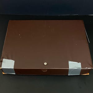 May include: A rectangular brown box with a metal finish. The box is sealed with grey tape at the corners and has a small, round metal clasp. The box is on a black surface.