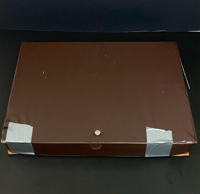 May include: A rectangular brown box with a metal finish. The box is sealed with grey tape at the corners and has a small, round metal clasp. The box is on a black surface.