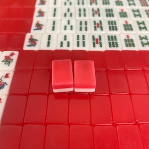 Puede incluir: Primer plano de un juego de Mahjong en curso, con fichas rojas y blancas. Dos fichas rojas con bases blancas est&aacute;n en foco, rodeadas de otras fichas con varios s&iacute;mbolos y caracteres. Las fichas est&aacute;n dispuestas sobre una superficie roja.