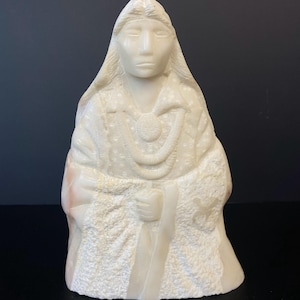 May include: A carved stone sculpture of a seated figure, likely a Native American woman, with long hair and a detailed robe. The sculpture is off-white with textured details and a necklace. The figure is holding an object in their right hand.