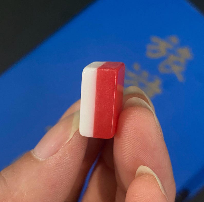 Puede incluir: Una peque&ntilde;a ficha de juego rectangular con una combinaci&oacute;n de colores blanco y rojo. La ficha se sostiene entre los dedos, con un fondo azul visible. El dise&ntilde;o de la ficha presenta una franja blanca vertical en un lado y una franja roja en el otro.