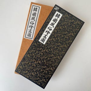 May include: Two rectangular boxes. One is a light brown color with a white label with black text. The other is black with a gold floral pattern and a white label with black text. The text on the labels is in a foreign language.