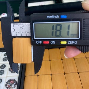 May include: A digital caliper measuring a light brown Mahjong tile. The tile is 18.1 mm wide. Other Mahjong tiles are visible in the background, along with a case.