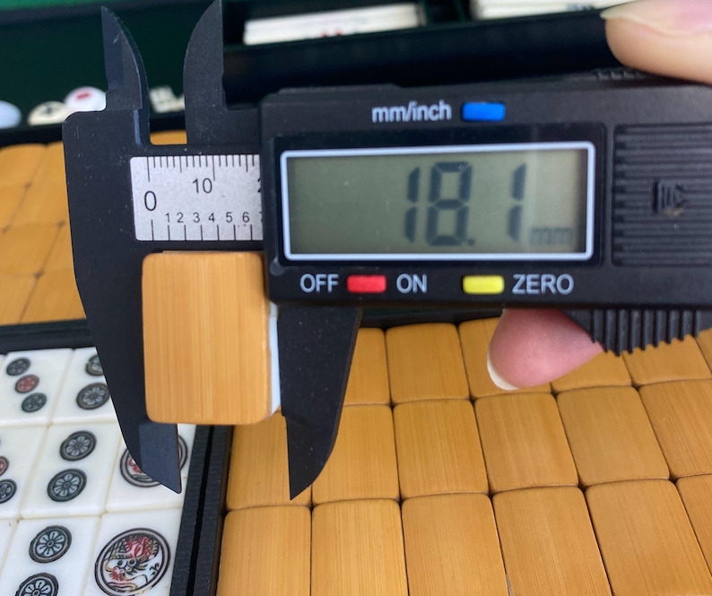 May include: A digital caliper measuring a light brown Mahjong tile. The tile is 18.1 mm wide. Other Mahjong tiles are visible in the background, along with a case.