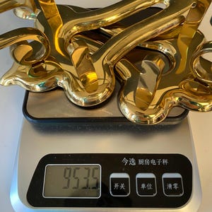 May include: A collection of shiny, gold-colored decorative hardware pieces, possibly door handles or drawer pulls, are stacked on a digital kitchen scale. The scale displays a weight of 953.5.