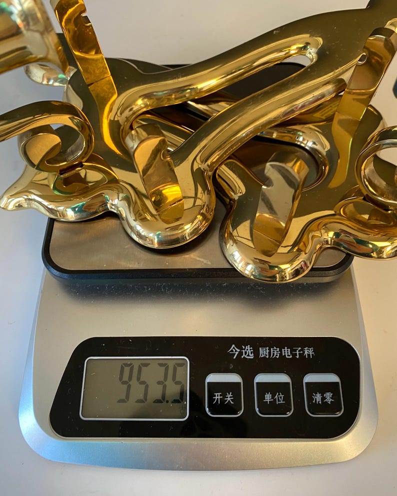May include: A collection of shiny, gold-colored decorative hardware pieces, possibly door handles or drawer pulls, are stacked on a digital kitchen scale. The scale displays a weight of 953.5.