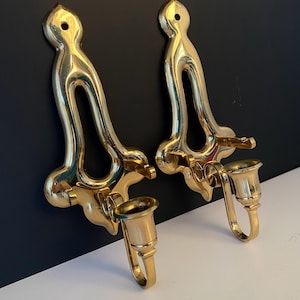 Puede incluir: Dos apliques de pared dorados y ornamentados. Cada aplique presenta un diseño curvo y abierto con un portavelas. Los apliques están montados sobre una superficie en blanco y negro, mostrando su elegante estilo vintage.