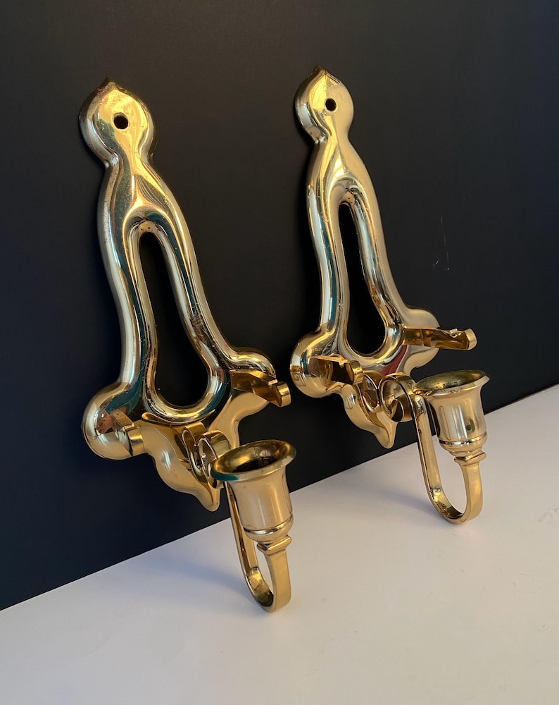 May include: Two ornate, gold-colored wall sconces. Each sconce features a curved, open design with a candle holder. The sconces are mounted on a black and white surface, showcasing their elegant, vintage style.