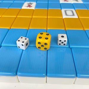 Puede incluir: Primer plano de una partida de Mahjong en curso. La imagen presenta fichas azules y amarillas, junto con tres dados. Un dado es amarillo y los otros dos son blancos. Cada dado tiene puntos negros que indican los n&uacute;meros.