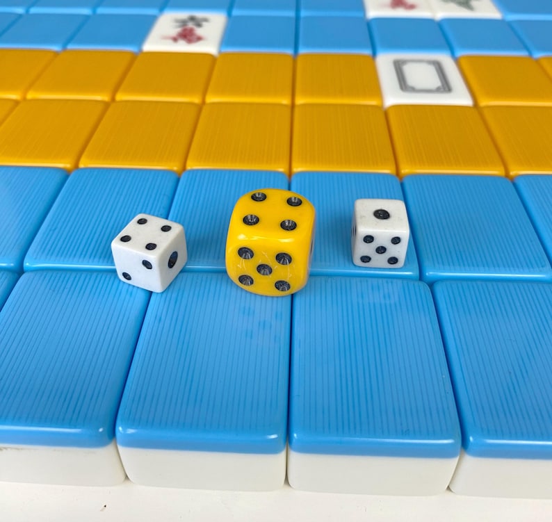 Puede incluir: Primer plano de una partida de Mahjong en curso. La imagen presenta fichas azules y amarillas, junto con tres dados. Un dado es amarillo y los otros dos son blancos. Cada dado tiene puntos negros que indican los n&uacute;meros.