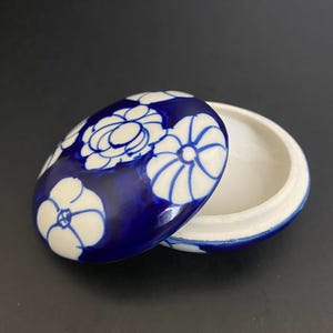 Op de afbeelding: Een klein, rond keramiek doosje met een deksel. Het deksel is versierd met witte bloemen op een diepblauwe achtergrond. De binnenkant van het doosje is wit.