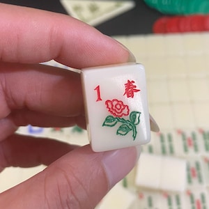 May include: A white Mahjong tile featuring the number 1 in red, a red rose with green leaves, and the Chinese character for "Spring" in red. The tile is held in a hand, with other tiles visible in the background.
