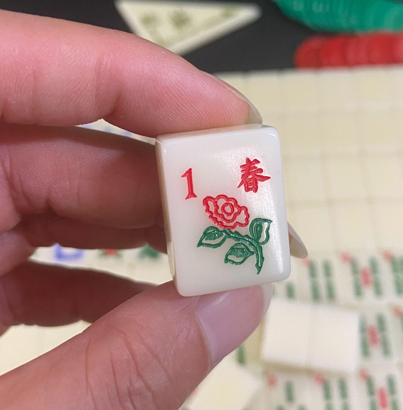 May include: A white Mahjong tile featuring the number 1 in red, a red rose with green leaves, and the Chinese character for "Spring" in red. The tile is held in a hand, with other tiles visible in the background.