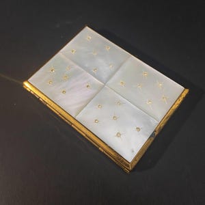 May include: A rectangular compact case featuring a gold-toned metal frame and four panels of iridescent white mother-of-pearl. Each panel is inlaid with small, star-shaped gold accents, creating a consistent pattern across the surface.