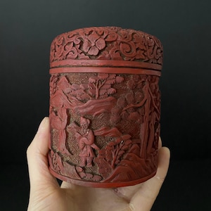 May include: A red cylindrical box with intricate carvings of a landscape scene with figures and trees. The box is made of lacquered wood and has a lid.