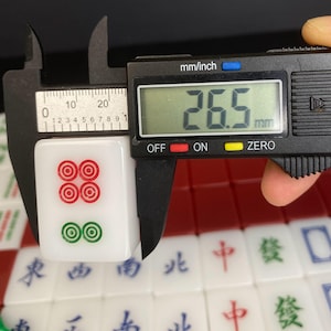 May include: A white mahjong tile with red and green circular symbols is measured with a digital caliper. The tile is 26.5 mm long. Other tiles with Chinese characters are visible in the background.