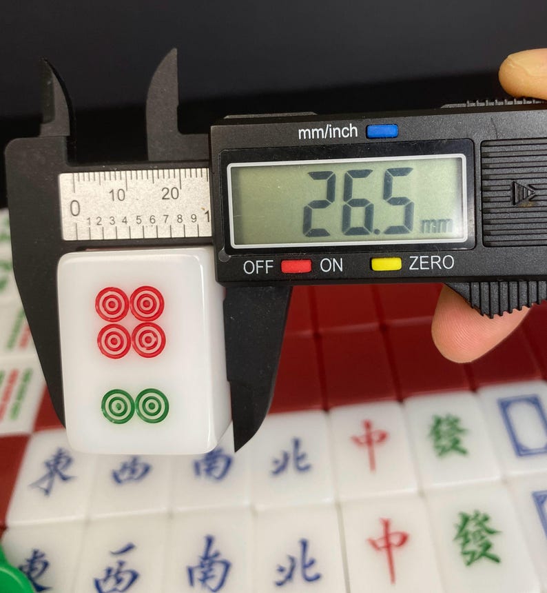May include: A white mahjong tile with red and green circular symbols is measured with a digital caliper. The tile is 26.5 mm long. Other tiles with Chinese characters are visible in the background.