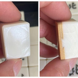 May include: Close-up of a white Mahjong tile with a smooth, glossy surface. The tile is held between fingers, showcasing its rectangular shape and the wooden side. Other tiles with Chinese characters are visible in the background.