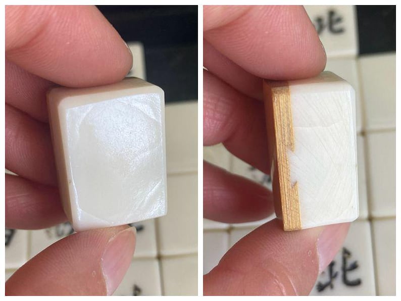 May include: Close-up of a white Mahjong tile with a smooth, glossy surface. The tile is held between fingers, showcasing its rectangular shape and the wooden side. Other tiles with Chinese characters are visible in the background.