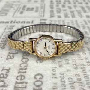 May include: A gold-tone wristwatch with a white face and black Roman numeral hour markers. The watch has a gold-tone band with a checkered pattern.