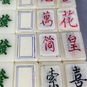 May include: Close-up of several off-white Mahjong tiles. Some tiles feature green or black Chinese characters, while others display red characters or blue-lined rectangles. The tiles are arranged in a grid, showcasing the variety of symbols and designs.