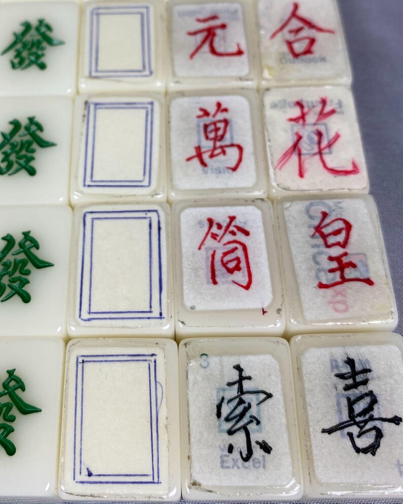 May include: Close-up of several off-white Mahjong tiles. Some tiles feature green or black Chinese characters, while others display red characters or blue-lined rectangles. The tiles are arranged in a grid, showcasing the variety of symbols and designs.