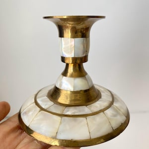 Pode incluir: Um castiçal de latão com uma base larga e um topo alargado. O corpo é adornado com azulejos de madrepérola embutidos, criando um padrão de mosaico. O metal tem um tom dourado quente.