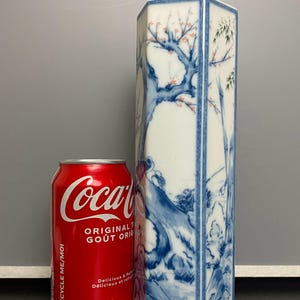 Puede incluir: Un jarrón de porcelana blanca con forma hexagonal y diseños florales azules y blancos. El jarrón está decorado con un árbol, bambú y otros motivos florales. El jarrón está al lado de una lata de Coca-Cola.