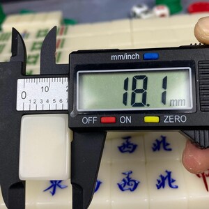 May include: A close-up of a white mahjong tile being measured with a digital caliper. The tile is 18.1 mm (0.71 inches) in length. The caliper displays the measurement in millimeters. Other mahjong tiles with blue characters are visible.