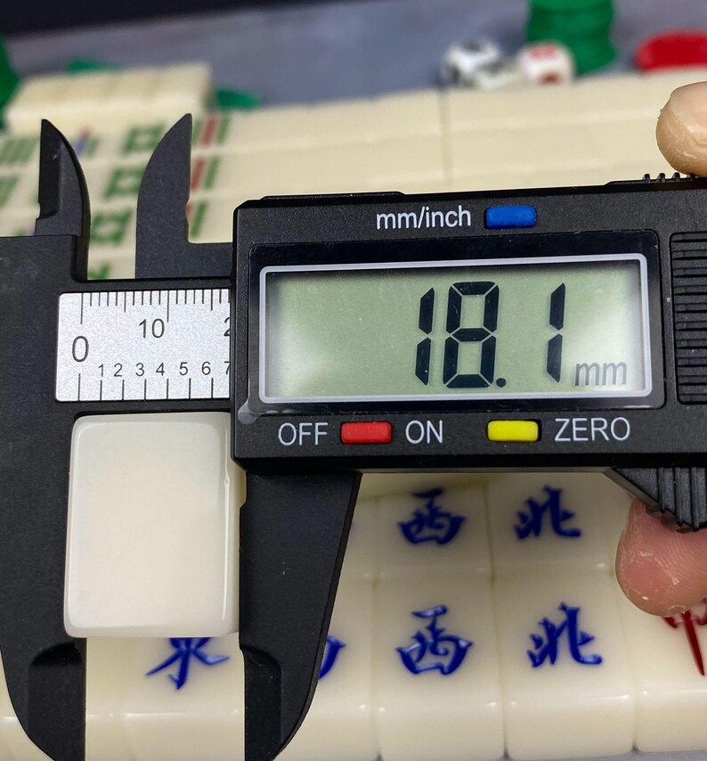 May include: A close-up of a white mahjong tile being measured with a digital caliper. The tile is 18.1 mm (0.71 inches) in length. The caliper displays the measurement in millimeters. Other mahjong tiles with blue characters are visible.